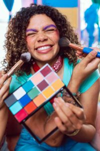 A cheerful woman with curly hair enjoys a colorful makeup session, smiling and holding a vibrant eyeshadow palette.